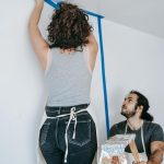 A couple working on a home renovation project using painter's tape and a ladder indoors.